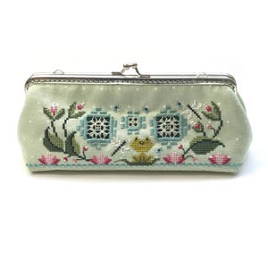Può includere: Una pochette in tessuto verde con una chiusura a scatto argentata. La pochette è decorata con un ricamo a punto croce che raffigura una rana seduta su una ninfea circondata da fiori e motivi geometrici.