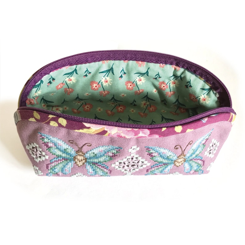 May include: A purple and green zippered pouch with a floral print and embroidered butterflies. The inside lining is a light green with a small white floral print.