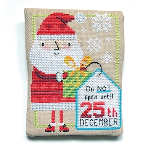 May include: A cross-stitched Christmas decoration featuring Santa Claus holding a wrapped gift. The tag reads "Do NOT open until 25th DECEMBER". The design includes snowflakes and is framed with a green border. The background is a beige fabric.