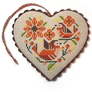 May include: A heart-shaped ornament featuring a cross-stitch design on beige fabric. The pattern displays two birds, orange flowers, and green leaves. The edge is finished with a dark brown scalloped trim and a braided cord for hanging.