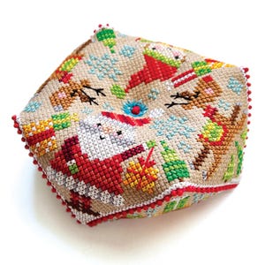 May include: A square, cross-stitched Christmas ornament featuring Santa Claus, reindeer, and snowflakes. The ornament is beige with red, green, white, and blue accents. The edges are trimmed with red pom-poms.