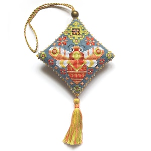 Può includere: Un ornamento a punto croce a forma di diamante raffigurante un'ape stilizzata con ali arancioni, gialle e bianche su sfondo blu. Il pezzo è decorato con motivi geometrici, un cordino intrecciato per appenderlo con una perlina e una nappa multicolore.