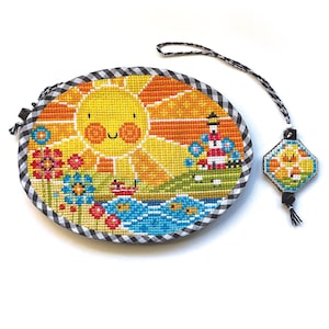 May include: Oval cross-stitch art featuring a smiling sun, flowers, a lighthouse, and a boat. The artwork is framed with a black and white checkered fabric. A matching charm with a tassel hangs from a black and white cord.
