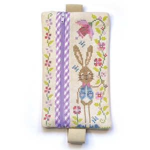 May include: A rectangular, beige cross-stitch pouch with a purple zipper and a beige loop. The pouch features a cross-stitched bunny in a blue vest, surrounded by flowers and butterflies. A purple and white checkered stripe runs down the center.