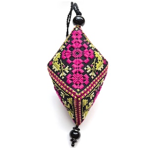 Puede incluir: Un adorno colgante en forma de diamante con intrincado bordado de punto de cruz sobre tela negra. El diseño muestra patrones florales geométricos en magenta vibrante y verde lima. Está rematado con un cordón negro y una cuenta negra, con una cuenta negra a juego en la punta inferior.