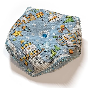 Può includere: Un cuscino decorativo quadrato, blu chiaro, ricamato a punto croce con una scena invernale. Il cuscino presenta un pupazzo di neve che tiene un cuore, insieme a stelle, alberi e altri elementi invernali. I bordi sono rifiniti con perline.