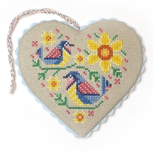 May include: Heart-shaped cross-stitch wall hanging featuring two blue and pink birds perched on a green vine with yellow flowers. The heart has a light blue scalloped edge and a braided hanging cord.