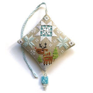 Pode incluir: Um enfeite de Natal bordado em bege e azul com uma rena, flocos de neve e um design de estrela. O enfeite tem uma conta azul e branca pendurada.