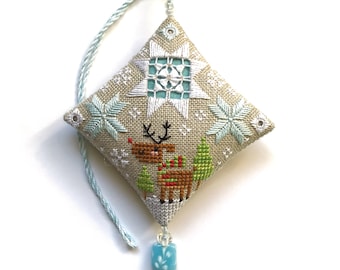 Puede incluir: Un adorno navideño bordado en beige y azul con un reno, copos de nieve y un diseño de estrella. El adorno tiene una cuenta azul y blanca que cuelga.