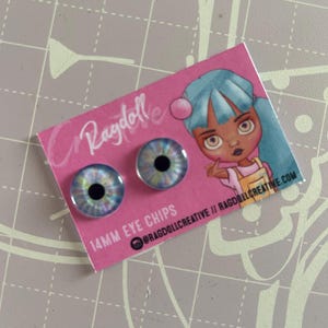 May include: Two 14mm eye chips with a colourful iris design, displayed on a pink card with a cartoon illustration and the text "Ragdoll" and "14MM EYE CHIPS". The eye chips are for crafts and doll making.