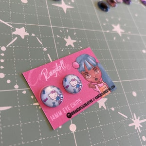 Puede incluir: Dos ojos redondos de 14 mm con un diseño de Hello Kitty. Los ojos están en una tarjeta rosa con el texto "14MM EYE CHIPS @RAGDOLLCREATIVE // RAGDOLLCREATIVE.COM".