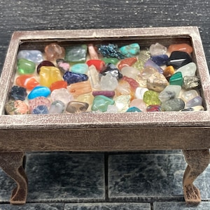 May include: A small wooden table with a glass top showcasing a collection of colourful gemstones. The table has a brown frame and decorative legs. The gemstones include red, blue, green, yellow, and clear stones.