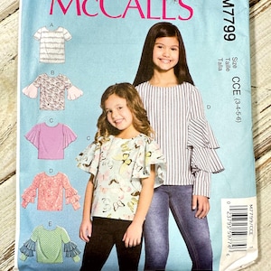 Può includere: Un modello di cucito McCall's, M7799, con illustrazioni di top da ragazza con diversi modelli di maniche. Il modello include vari stili e taglie CCE (3-4-5-6). La copertina è azzurra con il logo McCall's.