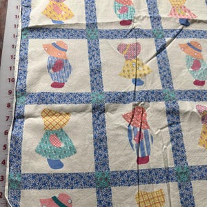 May include: White fabric with a blue and white border pattern. The fabric features a repeating pattern of patchwork dolls wearing dresses and hats in various colors.