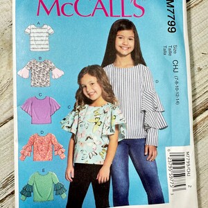 May include: McCall's sewing pattern M7799 featuring illustrations of girls' tops with various sleeve designs. The pattern includes sizes CHJ (7-8-10-12-14). The cover shows two girls wearing examples of the tops. Designed and printed in the USA.