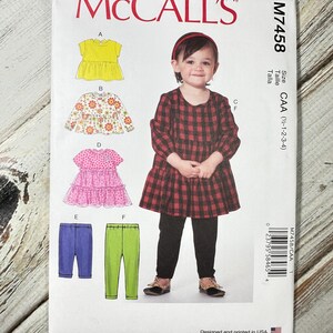 May include: McCall's sewing pattern M7458 featuring a young girl in a red and black plaid dress. The pattern includes illustrations of various tops and pants. The pattern is designed and printed in the USA.