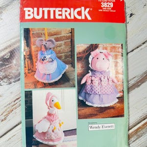 May include: A Butterick sewing pattern featuring three stuffed animal designs: a mouse, a pig, and a goose, each wearing a dress. The pattern cover is red and green with the Butterick logo and pattern number 3829.