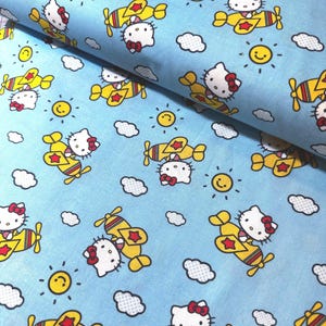 Może przedstawiać: Jasnoniebieska tkanina z powtarzającym się wzorem postaci Hello Kitty w żółtych samolotach, uśmiechniętych słońcach i białych chmurach. Odpowiednia do różnych projektów rzemieślniczych, w tym pikowania i odzieży.