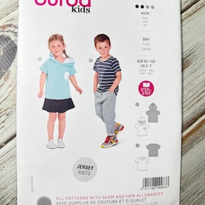 Puede incluir: Un paquete de patrones de costura Burda Kids con ilustraciones de ropa infantil. El paquete incluye una camisa con capucha azul, una camiseta a rayas y una falda. El texto en el paquete incluye "Jersey Knits" e información de tallas.