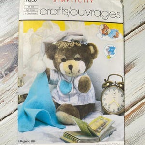 Puede incluir: Patrón de manualidades Simplicity con un oso de peluche con gorro de dormir y pijama. El patrón incluye el texto "Teddy Beddy Bear" y "Pattern Patron". También se ve un despertador y una manta azul.