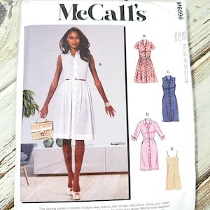 Op de afbeelding: Een McCall's naaipatroonpakket met een vrouw in een witte jurk. Het pakket bevat illustraties van verschillende jurkstijlen, waaronder een roze bloemenjurk, een spijkerjurk en een roze ruitjesjurk. De tekst op de verpakking luidt "McCall's".