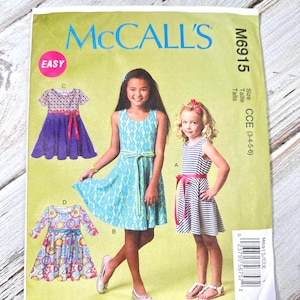 Può includere: Modello di cucito McCall's M6915 per abiti da ragazza. Il modello presenta illustrazioni di vari modelli di abiti, tra cui un abito in pizzo blu, un abito a righe con cintura rosa e un abito floreale. Il modello è etichettato "EASY".