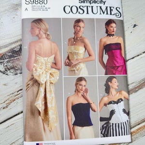 Peut inclure: Patron de couture Simplicity Costumes S9880, proposant des modèles de hauts sans bretelles. Tailles disponibles : U.S. 6-16, et tailles européennes 32-42. La couverture présente des idées de costumes, dont une robe dorée et une tenue rayée noire et blanche.
