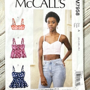 May include: A McCall's sewing pattern envelope featuring a woman wearing a crop top. The envelope displays illustrations of different crop top designs, including a pink gingham top. The pattern number is M7958.
