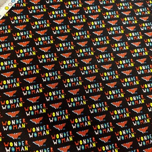 May include: Black fabric featuring a repeating pattern of the words "WONDER WOMAN" in white, yellow, blue, and red, along with the Wonder Woman logo. The fabric is suitable for various crafting projects.
