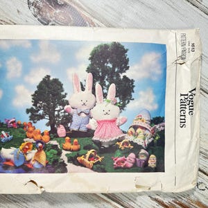 Könnte beinhalten: Vogue Patterns 1613. Das Schnittmustercover zeigt eine skurrile Osterszene mit Plüschhasen, Entenküken und dekorierten Eiern. Der Hintergrund zeigt einen blauen Himmel mit Wolken und Bäumen. Das Muster ist für eine Größe.