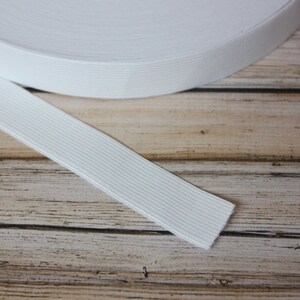 May include: White elastic band rolled up on a wooden surface. The elastic band is ribbed and has a smooth texture.