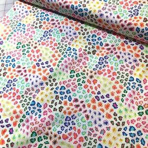 May include: White fabric with a colorful leopard print pattern. The pattern features small, irregular spots in shades of pink, orange, yellow, blue, and green. The fabric is folded, showing the pattern's repeating design.
