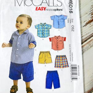 Può includere: Modello di cucito McCall's M6016 per camicie e pantaloncini per bambini. Il modello presenta vari stili di camicie in blu, rosso e a quadri, abbinati a pantaloncini gialli, a quadri e blu. Include una foto di un bambino con una camicia a quadri blu e bianca e pantaloncini blu.