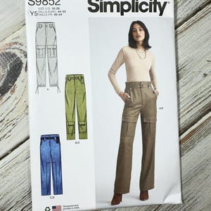 May include: Simplicity sewing pattern S9852 for cargo pants. The pattern includes illustrations of the pants in different colors and styles, with a model wearing a brown pair. The pattern size is U.S. 18-26.