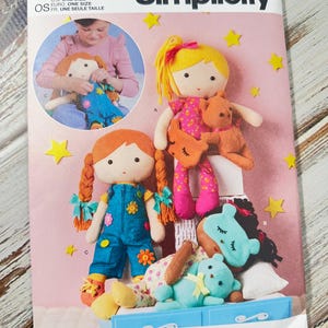 Pode incluir: Padrão de costura Simplicity com designs de bonecas. O padrão inclui instruções para criar bonecas de tecido com diferentes penteados e roupas. A capa mostra várias bonecas acabadas, incluindo uma com tranças laranjas.