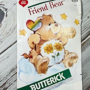 Könnte beinhalten: Ein Vintage-Butterick-Schnittmuster mit Friend Bear von Care Bears. Das Cover zeigt einen braunen Bären mit einem Regenbogenherz und einem mit Blumen geschmückten Bauch. Das Muster ist mit der Nummer 6231 und dem Care Bears-Logo gekennzeichnet.