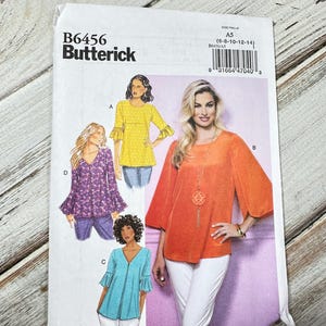 Pode incluir: Um padrão de costura Butterick, B6456, com ilustrações de tops femininos. O padrão inclui vários estilos, como um top amarelo com mangas com babados e um top laranja com mangas largas. A gama de tamanhos é A5 (6-8-10-12-14).