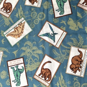 May include: A blue fabric with a repeating pattern of dinosaur illustrations. Each dinosaur is in a white frame with a brown border and the name of the dinosaur written below. The dinosaurs include Stegosaurus, Triceratops, Tyrannosaurus, Brontosaurus, and Pteranodon.
