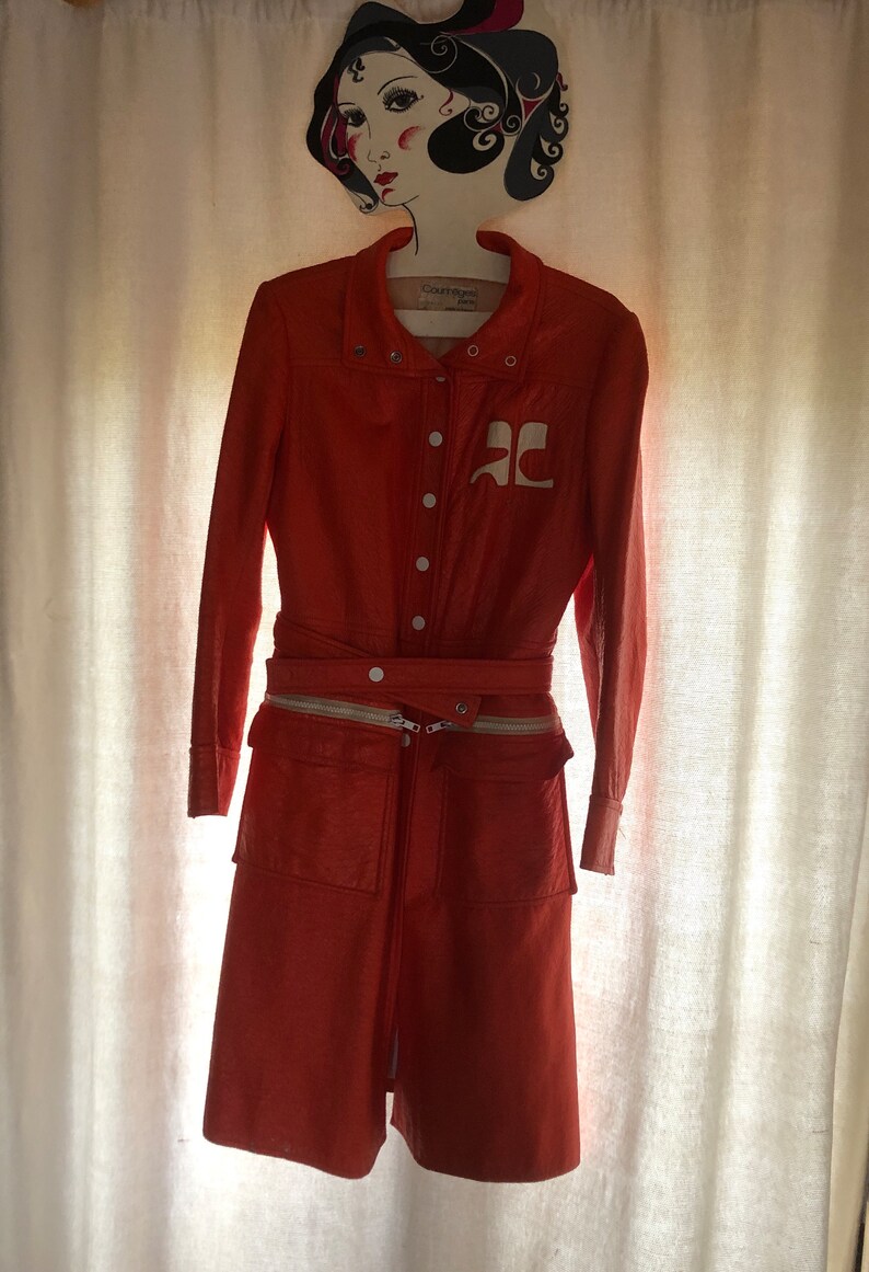 K&ouml;nnte beinhalten: Eine leuchtend orangefarbene Lederjacke mit einem wei&szlig;en Rei&szlig;verschluss und einem wei&szlig;en Logo auf der Vorderseite. Die Jacke hat einen G&uuml;rtel und zwei Taschen auf der Vorderseite.