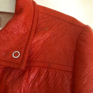K&ouml;nnte beinhalten: Nahaufnahme einer leuchtend orangefarbenen Lederjacke mit einem wei&szlig;en Patch auf der Vorderseite. Die Jacke hat einen Rei&szlig;verschluss und einen Kragen.