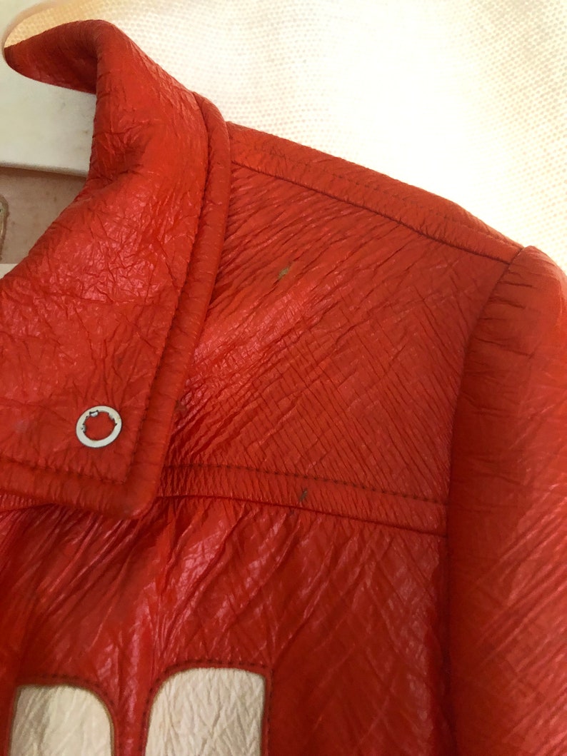 K&ouml;nnte beinhalten: Nahaufnahme einer leuchtend orangefarbenen Lederjacke mit einem wei&szlig;en Patch auf der Vorderseite. Die Jacke hat einen Rei&szlig;verschluss und einen Kragen.