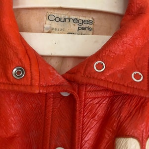 K&ouml;nnte beinhalten: Eine leuchtend orangefarbene Lederjacke mit einem wei&szlig;en Etikett, auf dem "Courreges Paris" und "08126" steht.