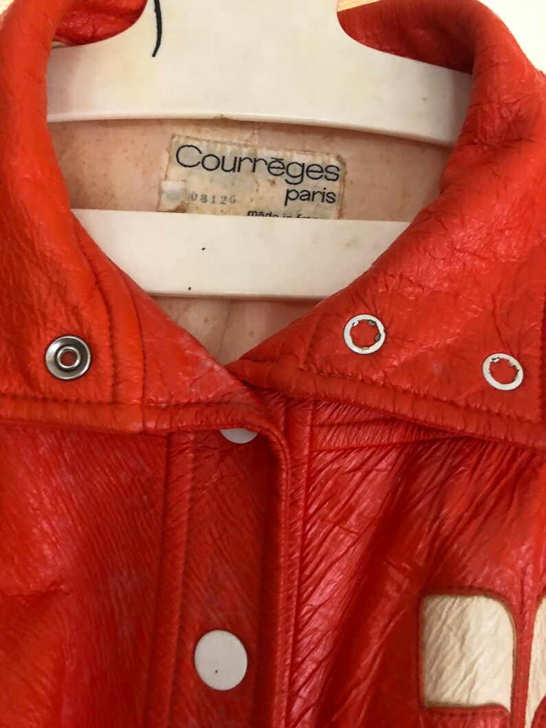 K&ouml;nnte beinhalten: Eine leuchtend orangefarbene Lederjacke mit einem wei&szlig;en Etikett, auf dem "Courreges Paris" und "08126" steht.