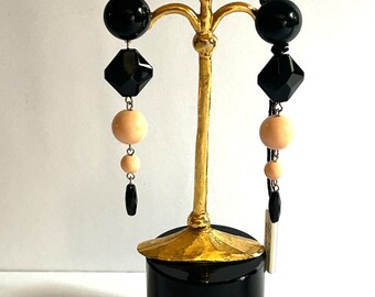 May include: Dangling earrings on a gold-coloured stand. Each earring has a black sphere, a black diamond-shaped bead, a peach-coloured sphere, a smaller peach-coloured sphere, and a black teardrop bead. The stand has a black base.
