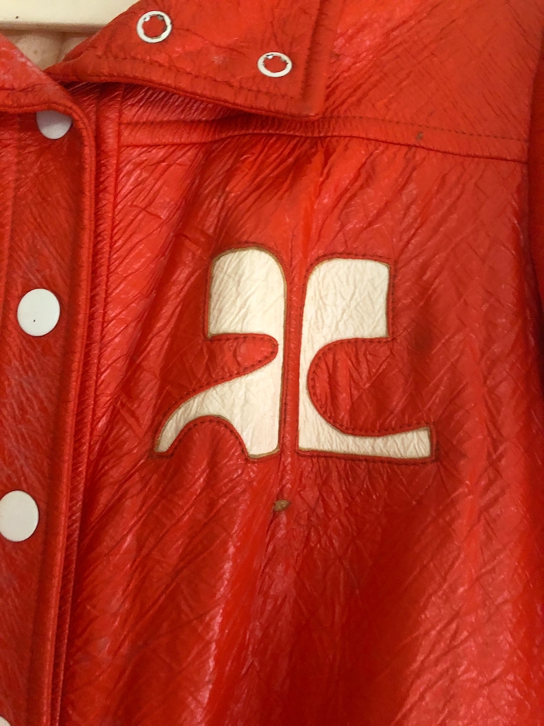 K&ouml;nnte beinhalten: Eine leuchtend orangefarbene Lederjacke mit einem wei&szlig;en Logo auf der Vorderseite. Das Logo ist ein stilisierter Buchstabe "A" mit einem geschwungenen Design.