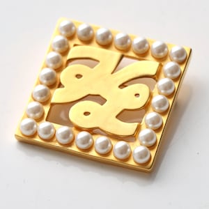 Peut inclure: Broche carrée dorée avec un motif ajouré central. La broche est bordée d'une rangée de fausses perles blanches et rondes. Le design est géométrique et élégant, adapté aux tenues de soirée ou pour ajouter une touche de glamour.