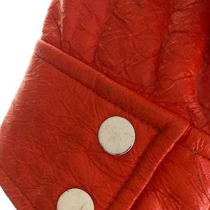 K&ouml;nnte beinhalten: Nahaufnahme einer leuchtend orangefarbenen Lederjacke mit wei&szlig;en Kn&ouml;pfen. Die Jacke hat eine strukturierte Oberfl&auml;che und eine Naht entlang der Manschette.