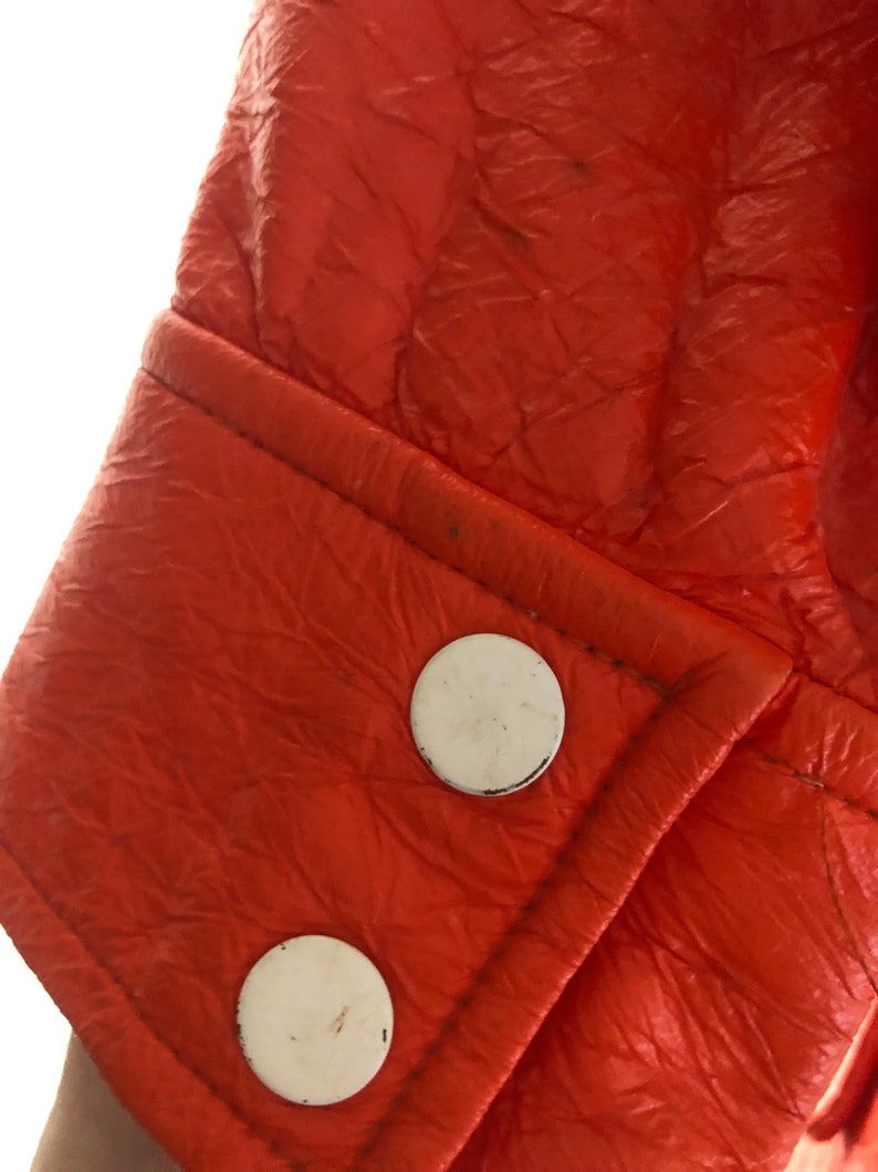 K&ouml;nnte beinhalten: Nahaufnahme einer leuchtend orangefarbenen Lederjacke mit wei&szlig;en Kn&ouml;pfen. Die Jacke hat eine strukturierte Oberfl&auml;che und eine Naht entlang der Manschette.