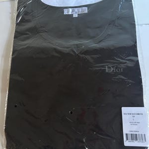 May include: Black t-shirt with a white Dior logo on the front. The shirt is packaged in a clear plastic bag with a white tag that reads "Beauty Worn Black Animation Top" and "Dior Homme".