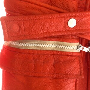 K&ouml;nnte beinhalten: Nahaufnahme einer leuchtend orangefarbenen Lederjacke mit Rei&szlig;verschluss und wei&szlig;en Kn&ouml;pfen. Die Jacke hat eine strukturierte Oberfl&auml;che und einen wei&szlig;en Rei&szlig;verschluss.
